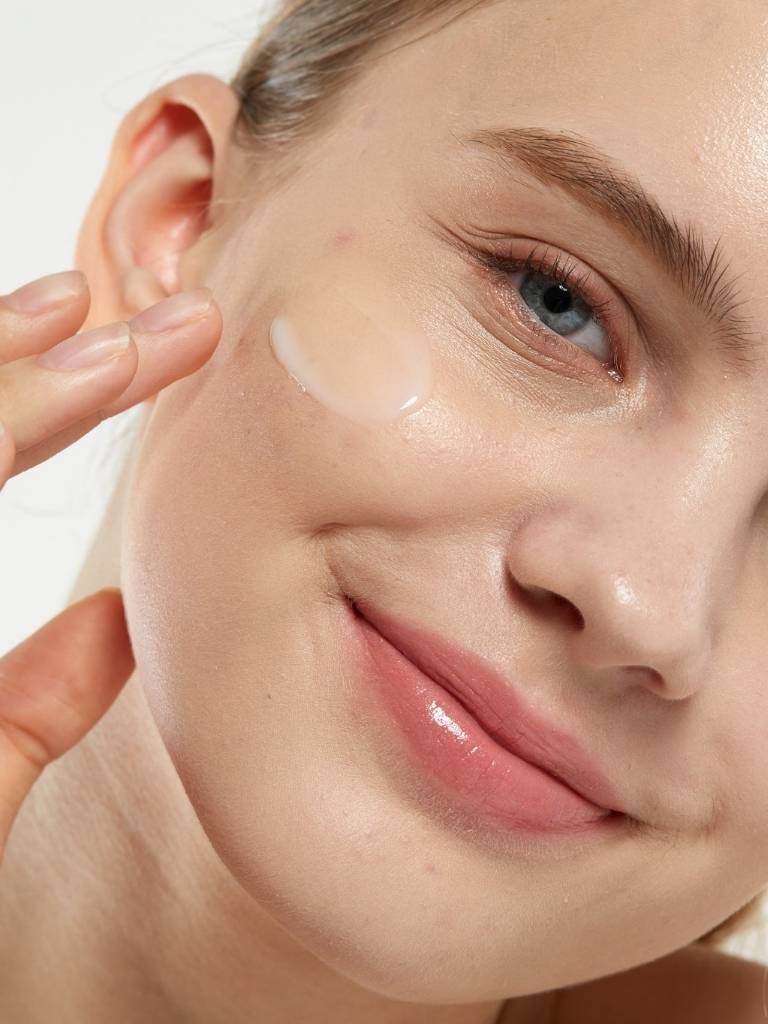 Close-up of a woman applying cream to her face with a neutral background