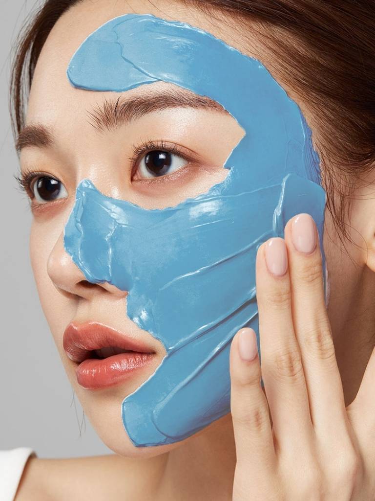 Woman applying blue facial mask to her face against a neutral background