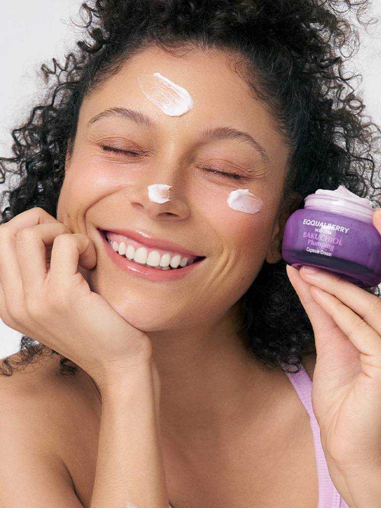 Woman applying cream to her face with a purple jar of cream next to her.