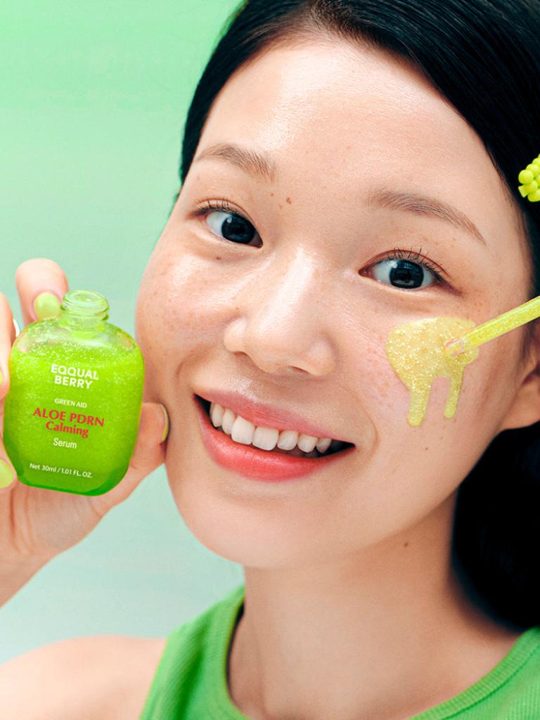 Woman applying aloe vera gel to her face with a green serum bottle in hand on a light green background