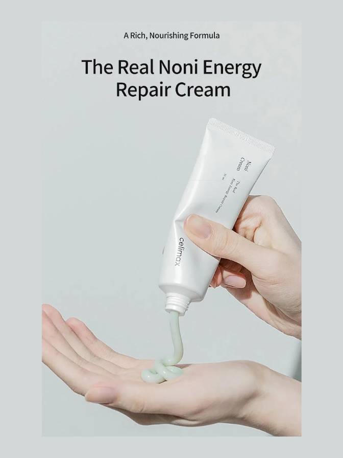 Hand holding a tube of 'The Real Noni Energy Repair Cream' with cream being squeezed onto a palm against a light gray background.